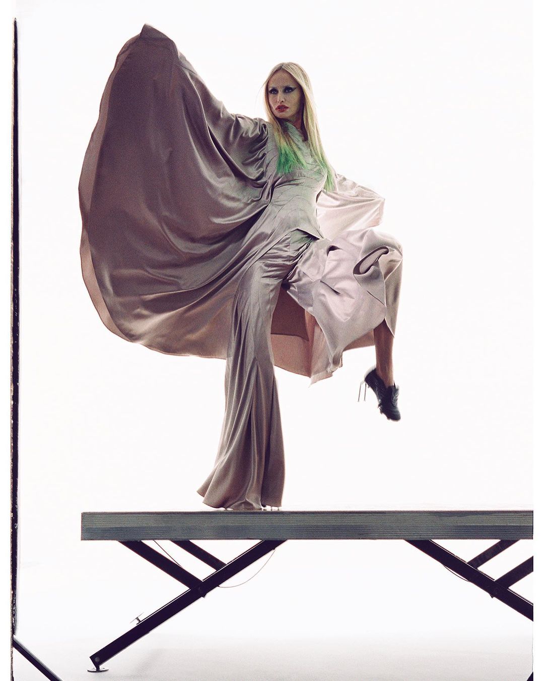 Fashion shot of a woman in a long gown, elevated on a platform with white backdrop, showcasing avant-garde style and dramatic pose.