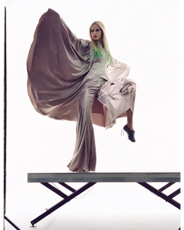 Fashion shot of a woman in a long gown, elevated on a platform with white backdrop, showcasing avant-garde style and dramatic pose.