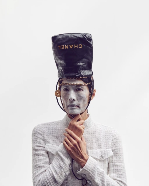 Woman with face paint and a Chanel handbag on her head in a fashion editorial, wearing a white tweed jacket against a plain white backdrop.