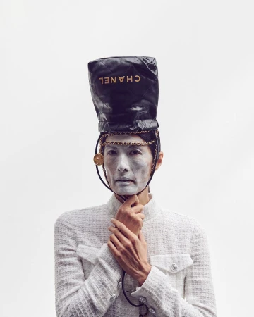 Woman with face paint and a Chanel handbag on her head in a fashion editorial, wearing a white tweed jacket against a plain white backdrop.