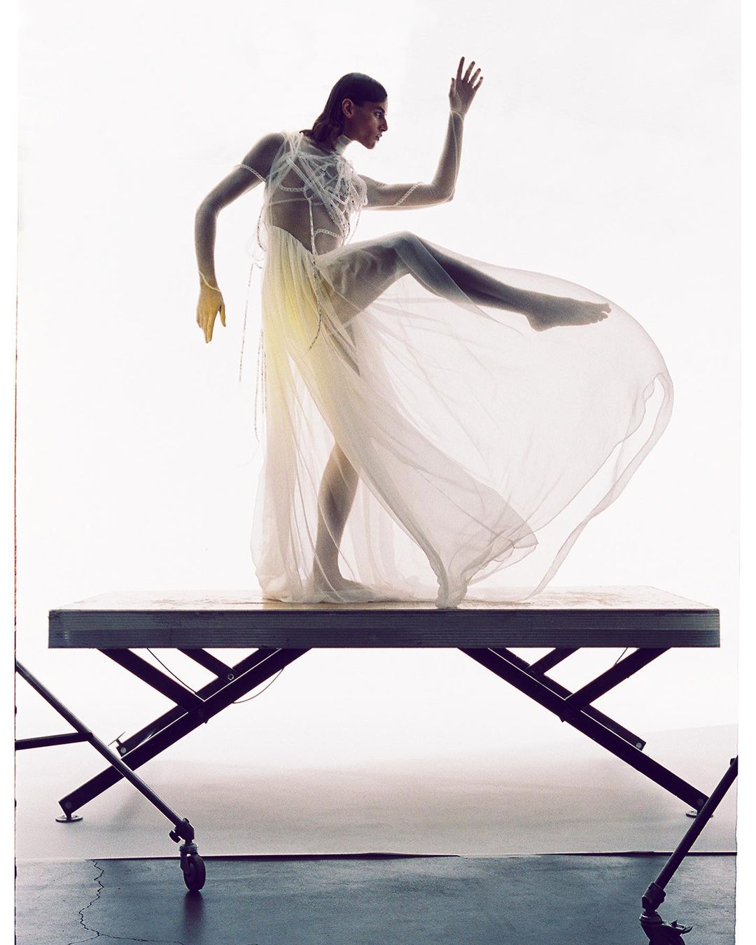 Woman in a sheer dress strikes a dynamic pose on a metal table against a bright background, showcasing fashion and artistic expression.