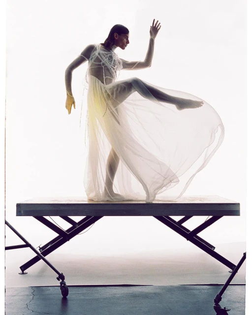 Woman in a sheer dress strikes a dynamic pose on a metal table against a bright background, showcasing fashion and artistic expression.