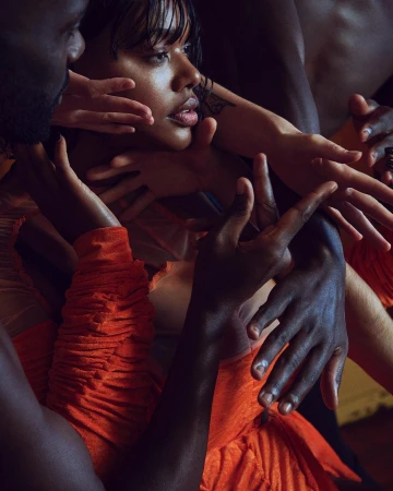 Close-up portrait of a woman in an orange dress surrounded by multiple hands touching her face and shoulders