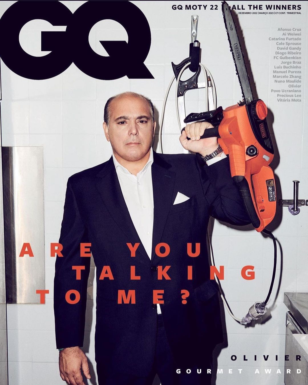 GQ magazine cover featuring a man in a suit holding a chainsaw in a kitchen setting, with bold typographic elements and GQ branding.