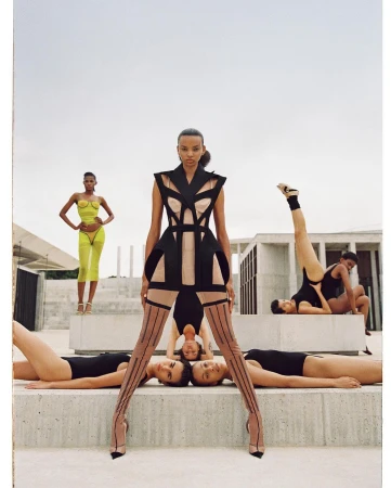 Fashion editorial featuring a woman in a structured black dress standing among a group of women on a concrete rooftop against a sky background.