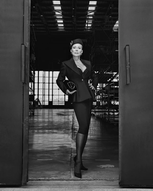 Elegant woman in vintage-inspired attire poses in a hangar doorway, holding a purse, with industrial scaffolding in the background, in black and white.