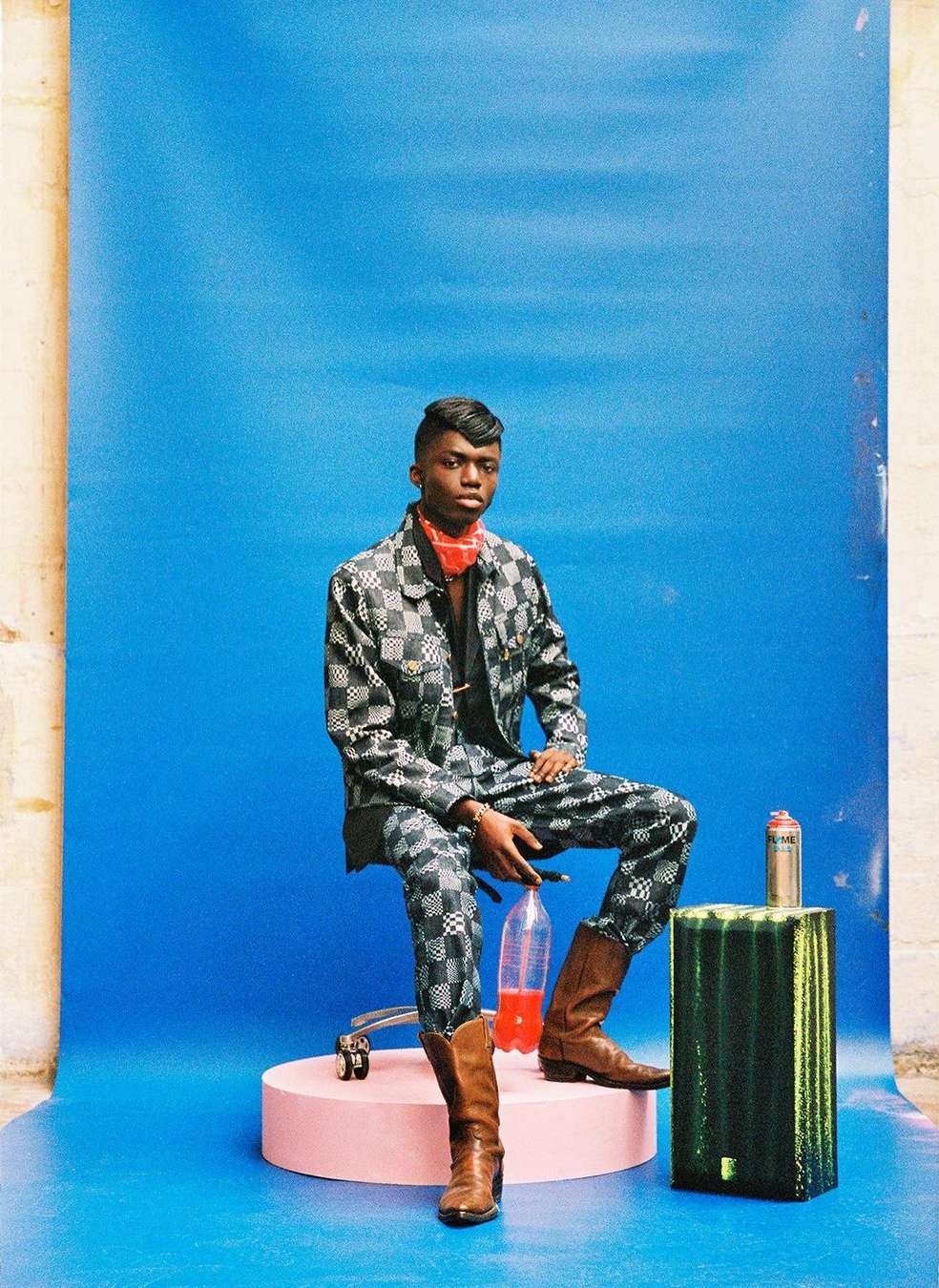 A young man in a checkered suit poses seated against a blue backdrop with various props. He is wearing brown boots and a red bandana.