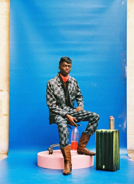 A young man in a checkered suit poses seated against a blue backdrop with various props. He is wearing brown boots and a red bandana.