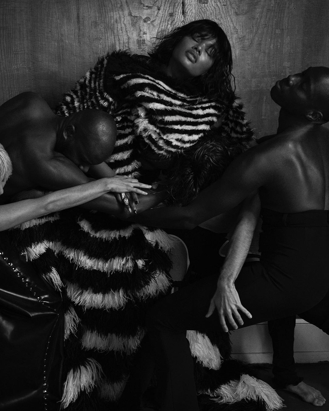 Black and white image of a woman in a zebra-striped garment surrounded by two bald men in a studio setting, portrait orientation.