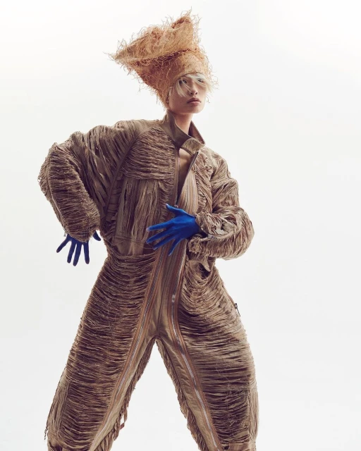 Model poses in a textured tan jumpsuit with blue gloves and a straw headpiece against a white backdrop in a studio fashion shot.