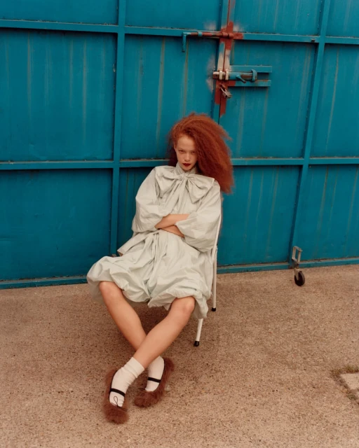 A young woman with red hair poses in a light green dress and furry shoes against a blue metal door backdrop. The shot is a medium, full-body capture.