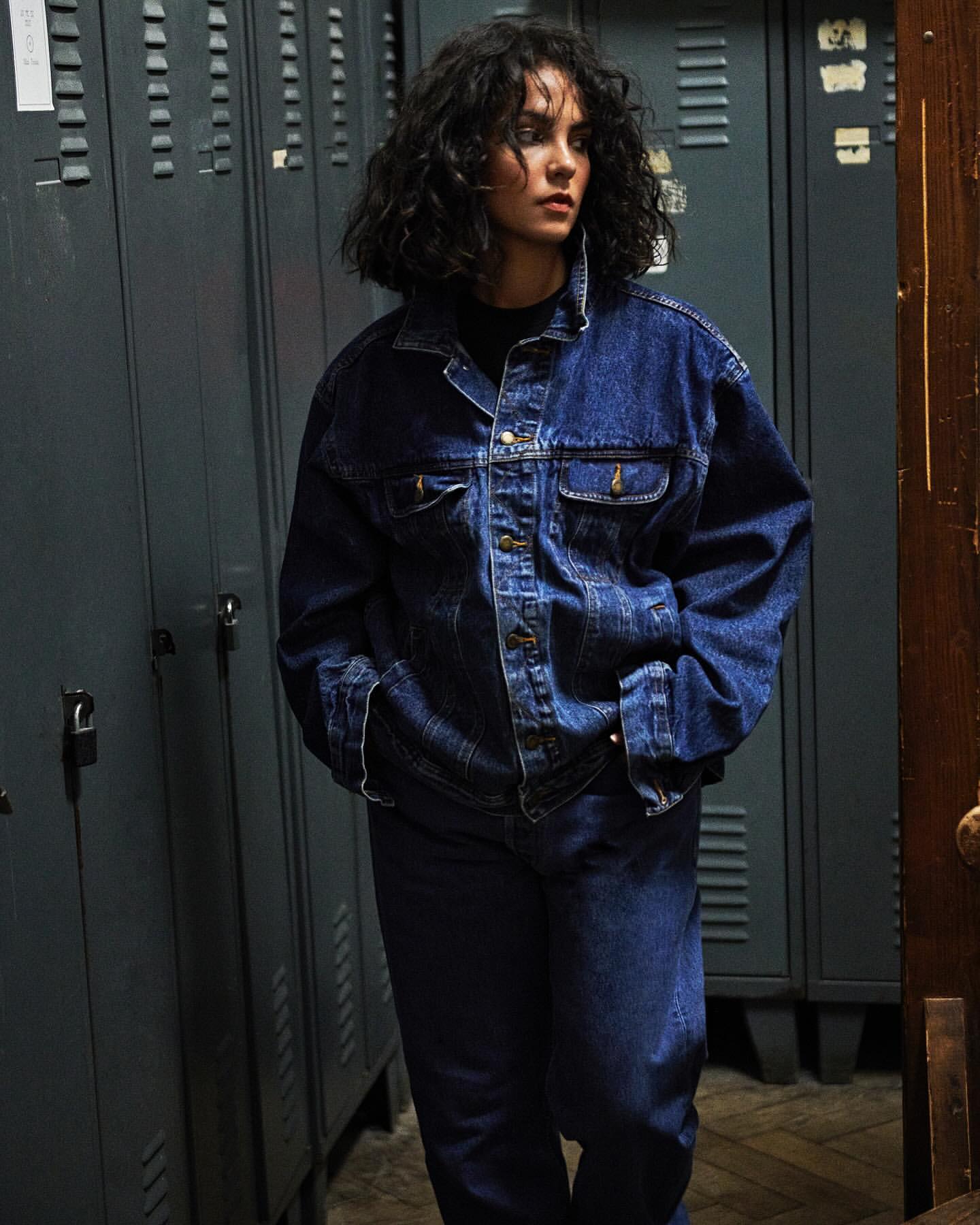Young woman with curly hair poses in a denim jacket and jeans next to gray metal lockers and a dark wood door frame indoors.
