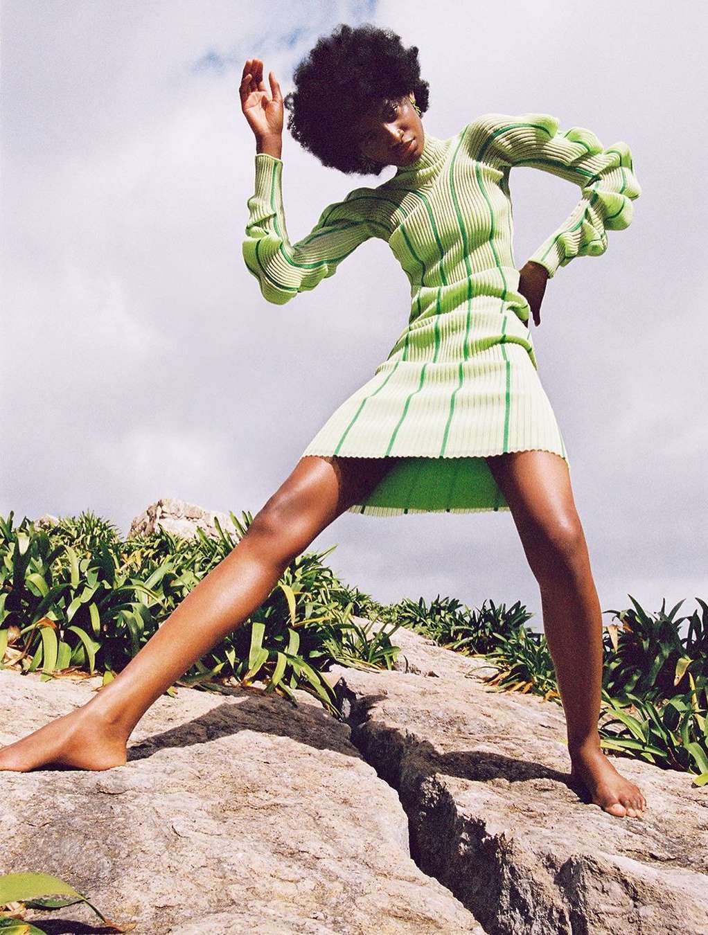 Fashionable black female model in a green ribbed dress standing on rocks outdoors against a partly cloudy sky in a dynamic pose.