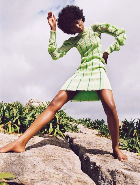 Fashionable black female model in a green ribbed dress standing on rocks outdoors against a partly cloudy sky in a dynamic pose.