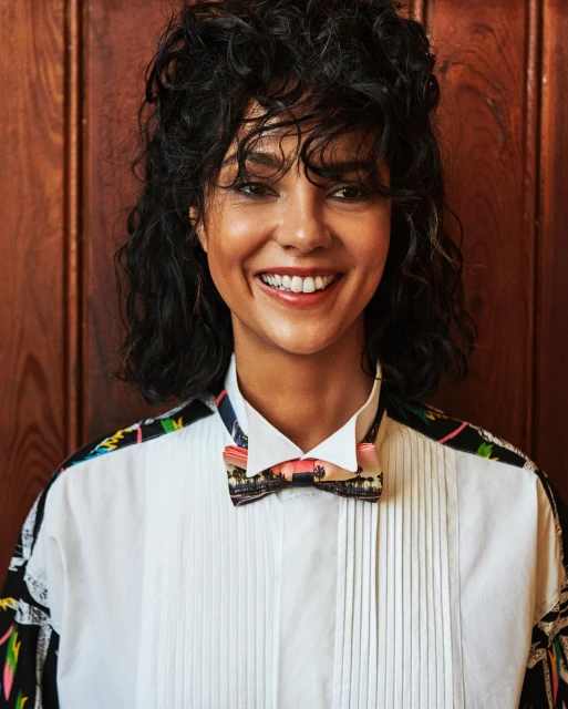 A smiling woman with short, curly hair wearing a white shirt with patterned sleeves and a landscape-printed bow tie, standing indoors.