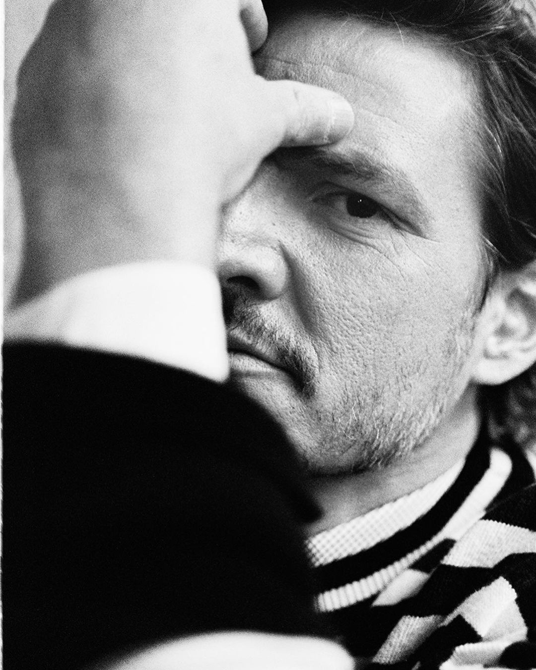 Black and white portrait of an adult male with a mustache, partially obscured by a hand over his face, wearing a striped sweater.