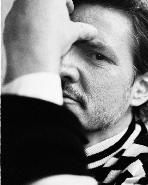 Black and white portrait of an adult male with a mustache, partially obscured by a hand over his face, wearing a striped sweater.