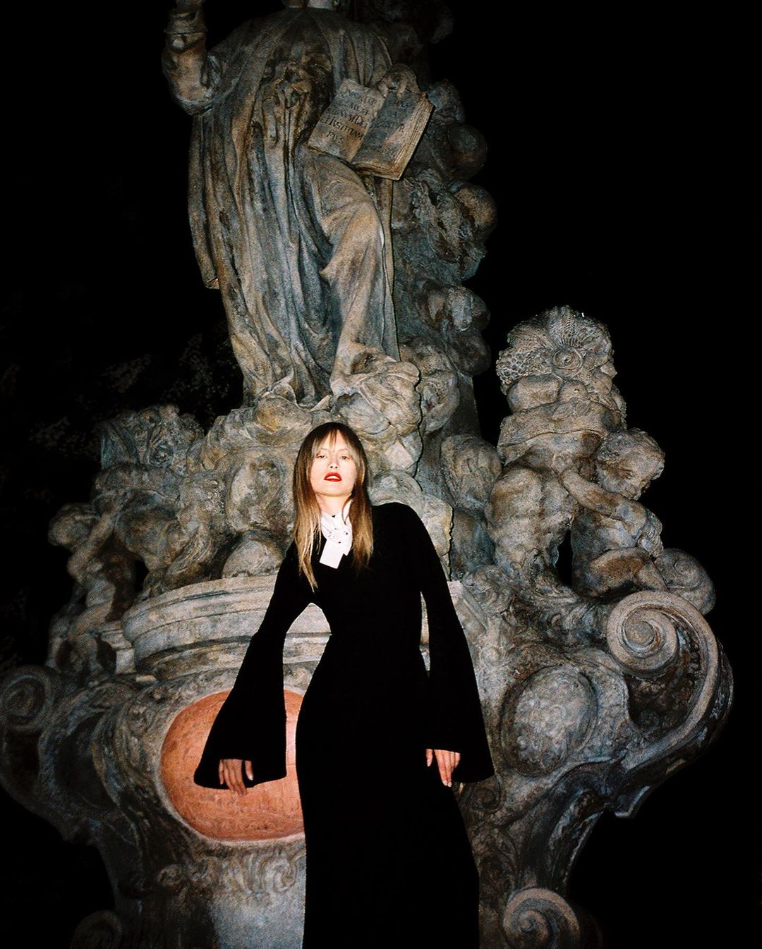 A female model stands in front of an old marble statue at night, wearing a floor-length black dress with long sleeves and a high collar.