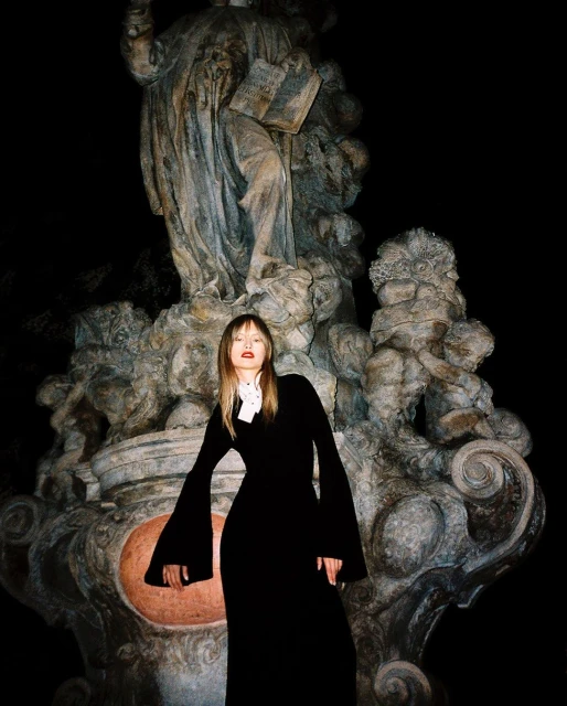 A female model stands in front of an old marble statue at night, wearing a floor-length black dress with long sleeves and a high collar.