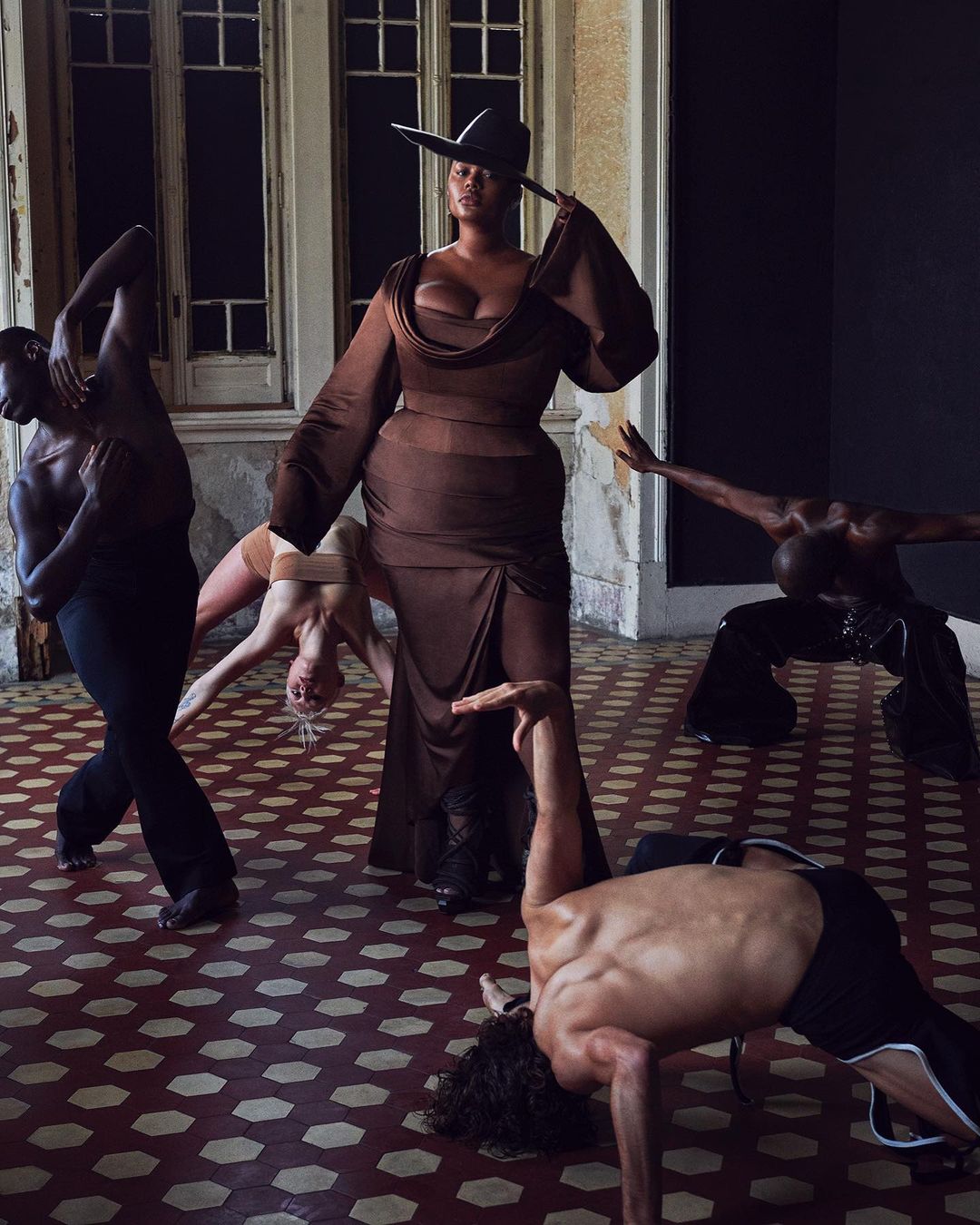 A woman in a brown dress and hat stands centrally amidst five dancers in a large room with tiled floors and large windows.