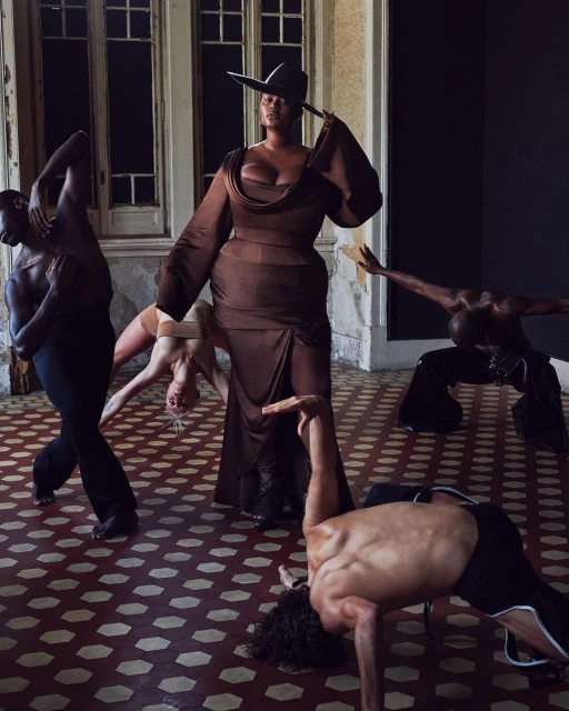 A woman in a brown dress and hat stands centrally amidst five dancers in a large room with tiled floors and large windows.