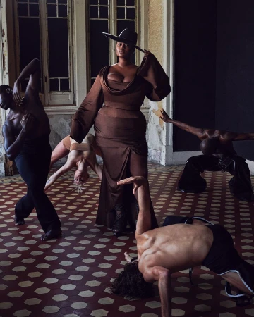 A woman in a brown dress and hat stands centrally amidst five dancers in a large room with tiled floors and large windows.
