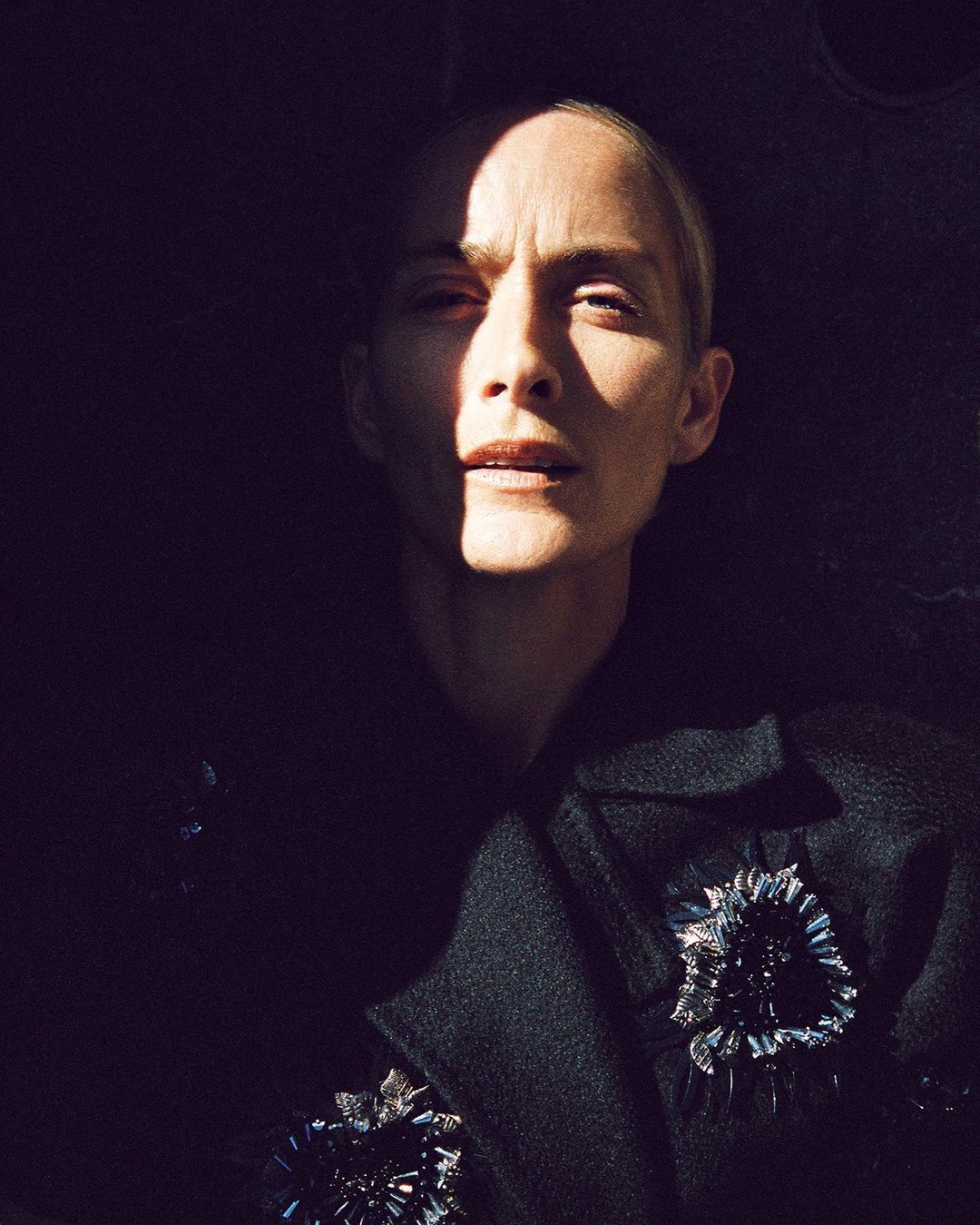 Close-up studio portrait of an adult woman in a black jacket with decorative brooches under dramatic lighting, creating a high-contrast visual.