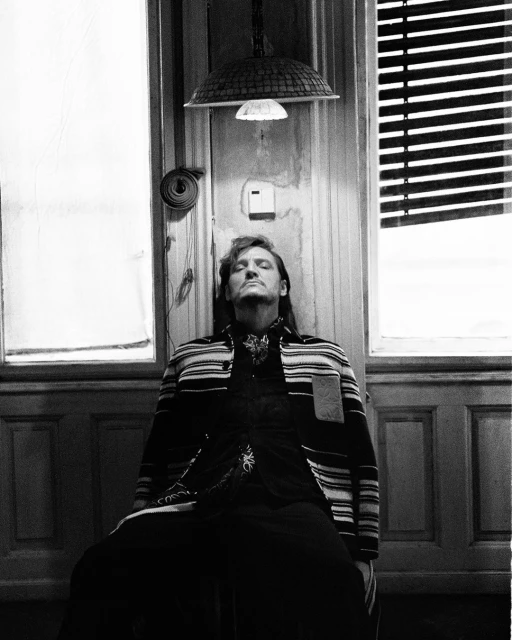 Black and white portrait of a man with long hair, sitting against a paneled wall with closed eyes, illuminated by a lamp.