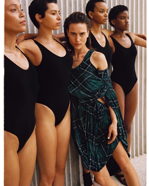Group of five women in black leotards and a green plaid dress posing against a wall, fashion-forward aesthetic with diverse representation.