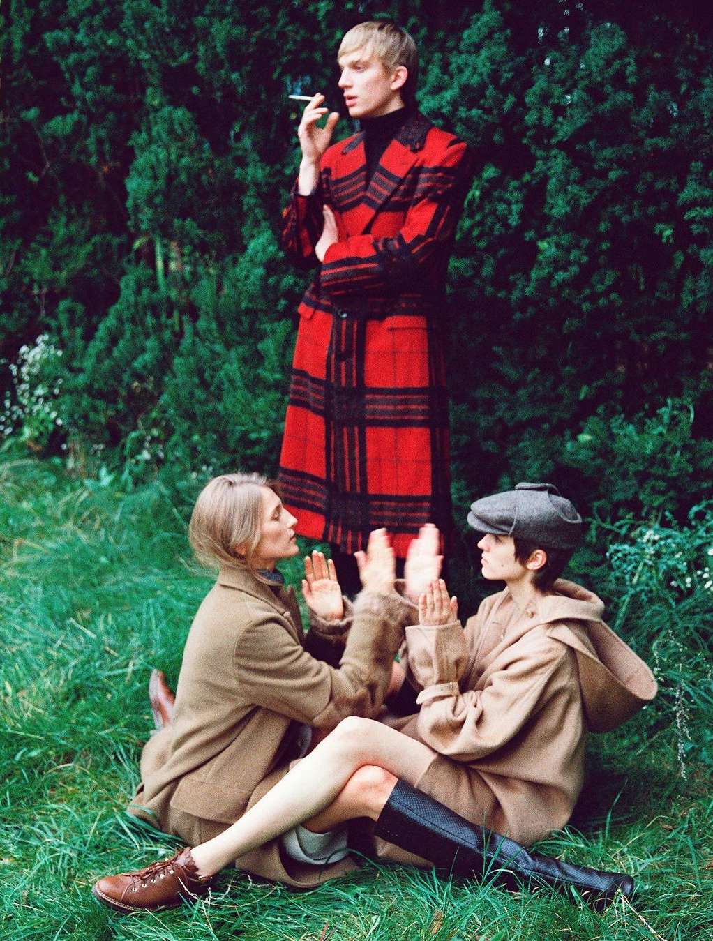Three models in a grassy outdoor setting: two women sitting on the ground, one man standing behind smoking a cigarette.