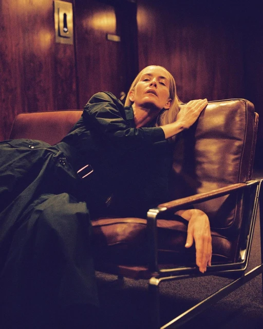 Adult woman with blonde hair reclines in a brown leather chair in an interior setting with wood paneling, wearing a dark dress.