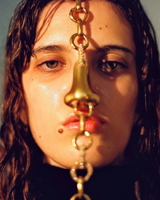Close-up portrait of a woman with golden nose jewelry and dark hair, shot at eye-level in a photo studio with soft lighting.