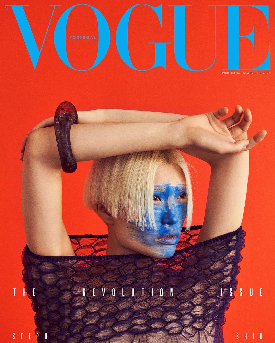 Vogue Portugal magazine cover featuring a woman with blonde hair and blue paint on her face, set against an orange background.
