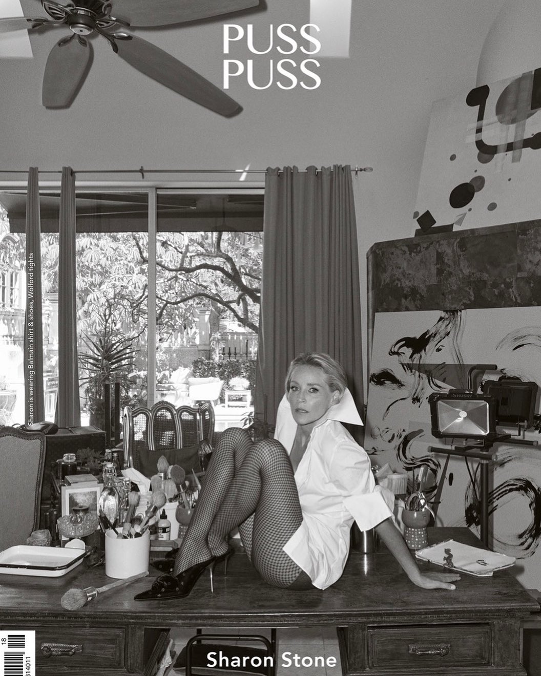 Black and white shot of Sharon Stone sitting on a desk in fishnets and an oversized white shirt, looking at the camera with an art studio backdrop.
