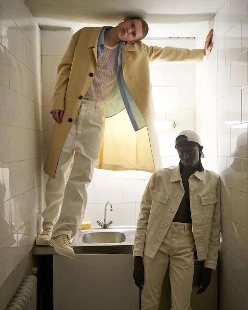 Two men pose indoors in matching neutral-tone outfits, one standing on a bathroom counter in a tan overcoat for a fashion editorial.