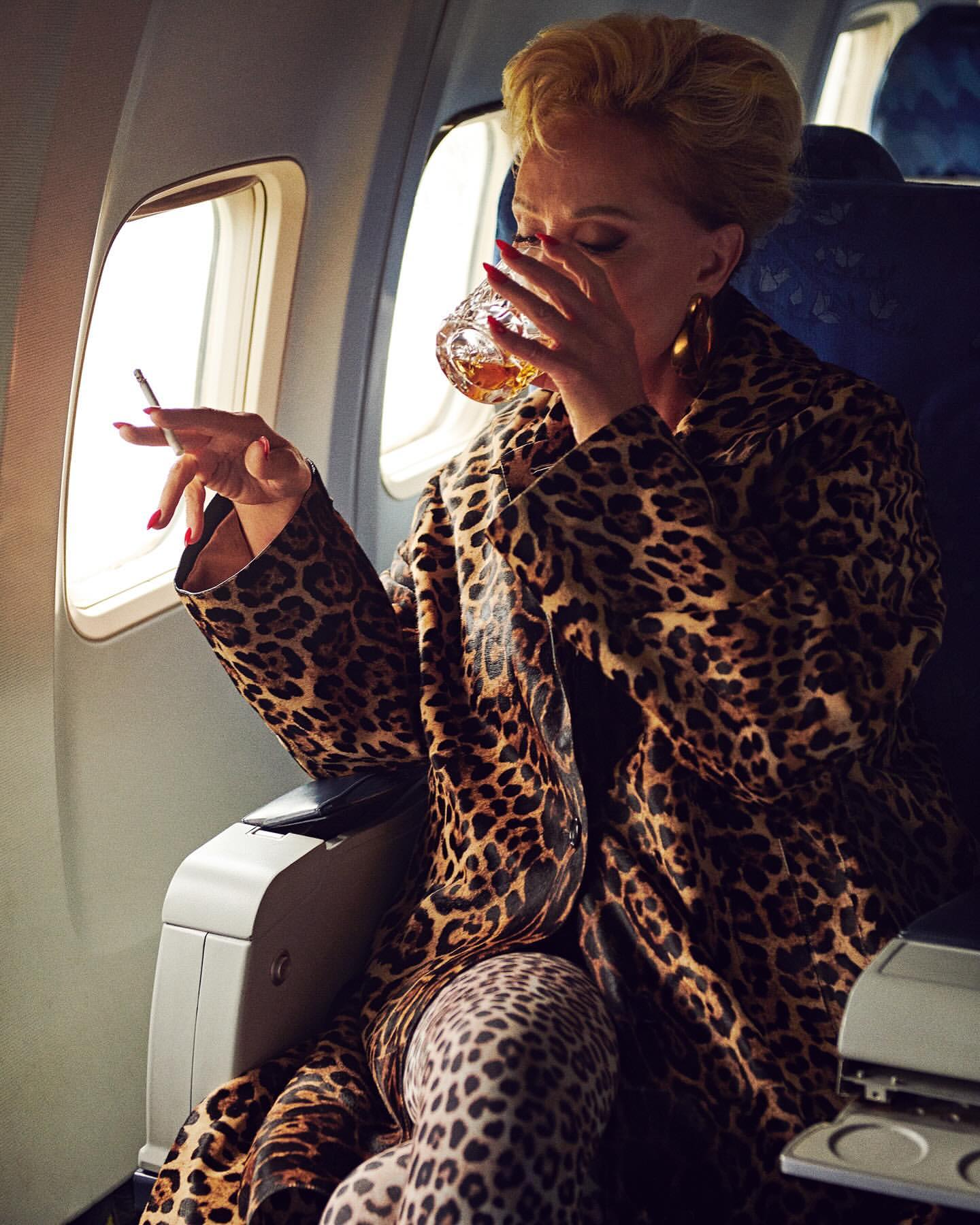 Adult female in airplane seat wearing leopard print coat, drinking from a glass, holding a cigarette and sitting by a window.