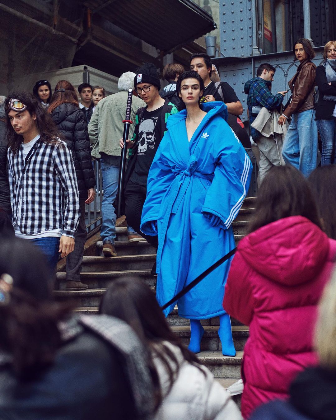 Woman in oversized blue Adidas robe and matching boots walking in a crowd, fashion editorial shot in an urban setting on steps.