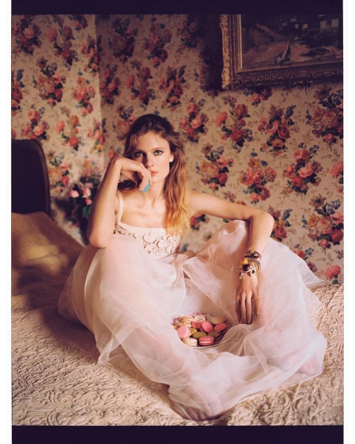 Woman in a gown sits on a bed with macarons in her lap, posing in a room with floral wallpaper and a framed painting.