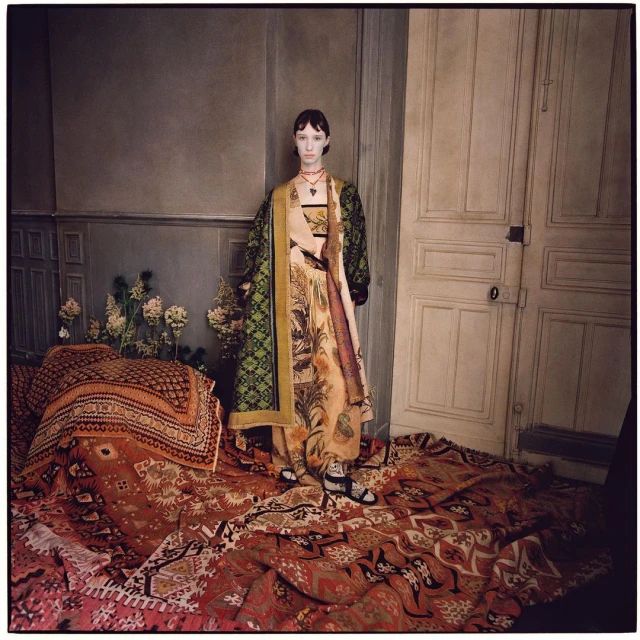 A fashion photo features a slender woman in patterned clothing standing on a rug indoors with patterned textiles and flowers nearby.
