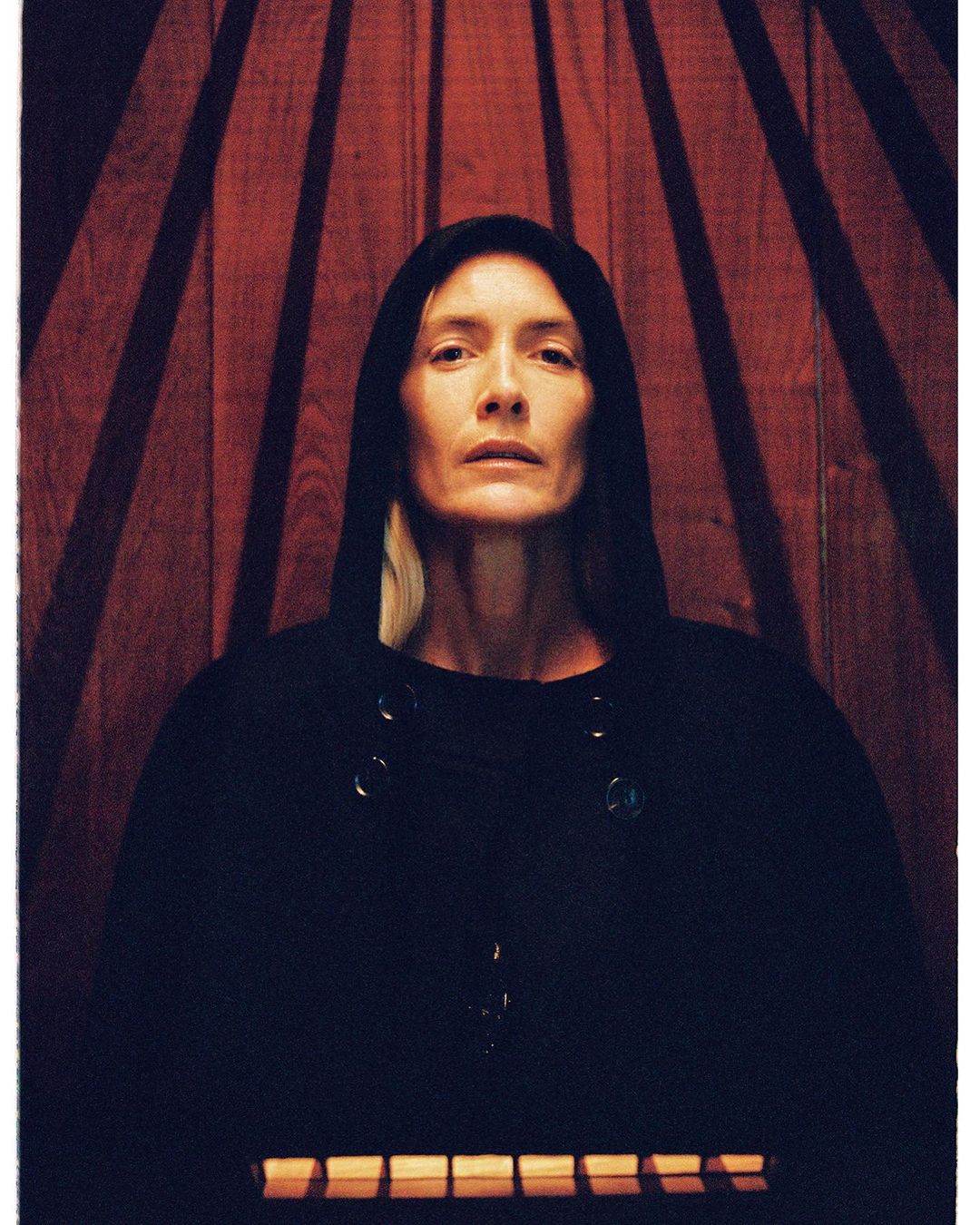 Portrait of a mature adult woman wearing a hooded top, against a wood-panelled wall with striped lighting effects.