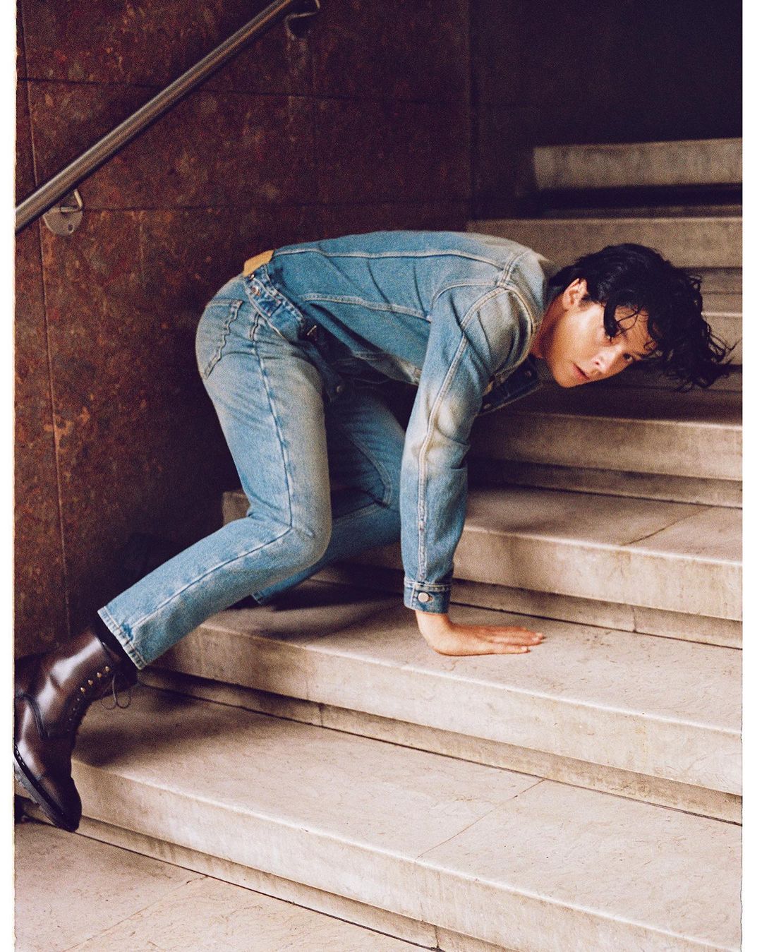 An adult crawls on marble stairs wearing a light blue denim jacket and jeans with black leather boots in an indoor setting.