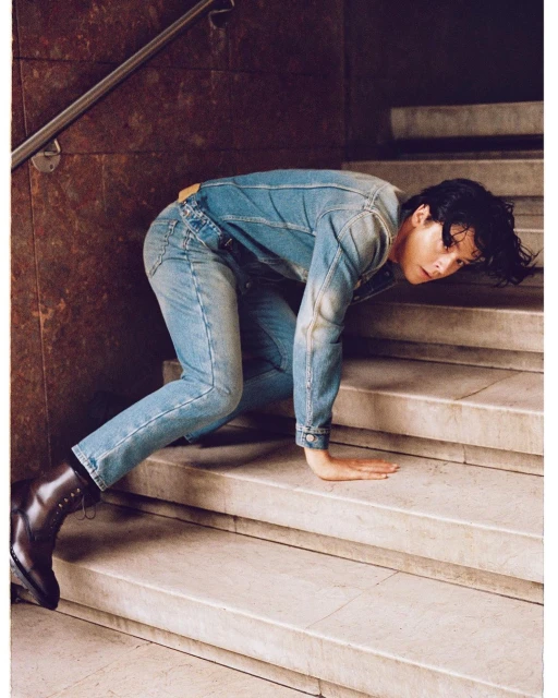 An adult crawls on marble stairs wearing a light blue denim jacket and jeans with black leather boots in an indoor setting.