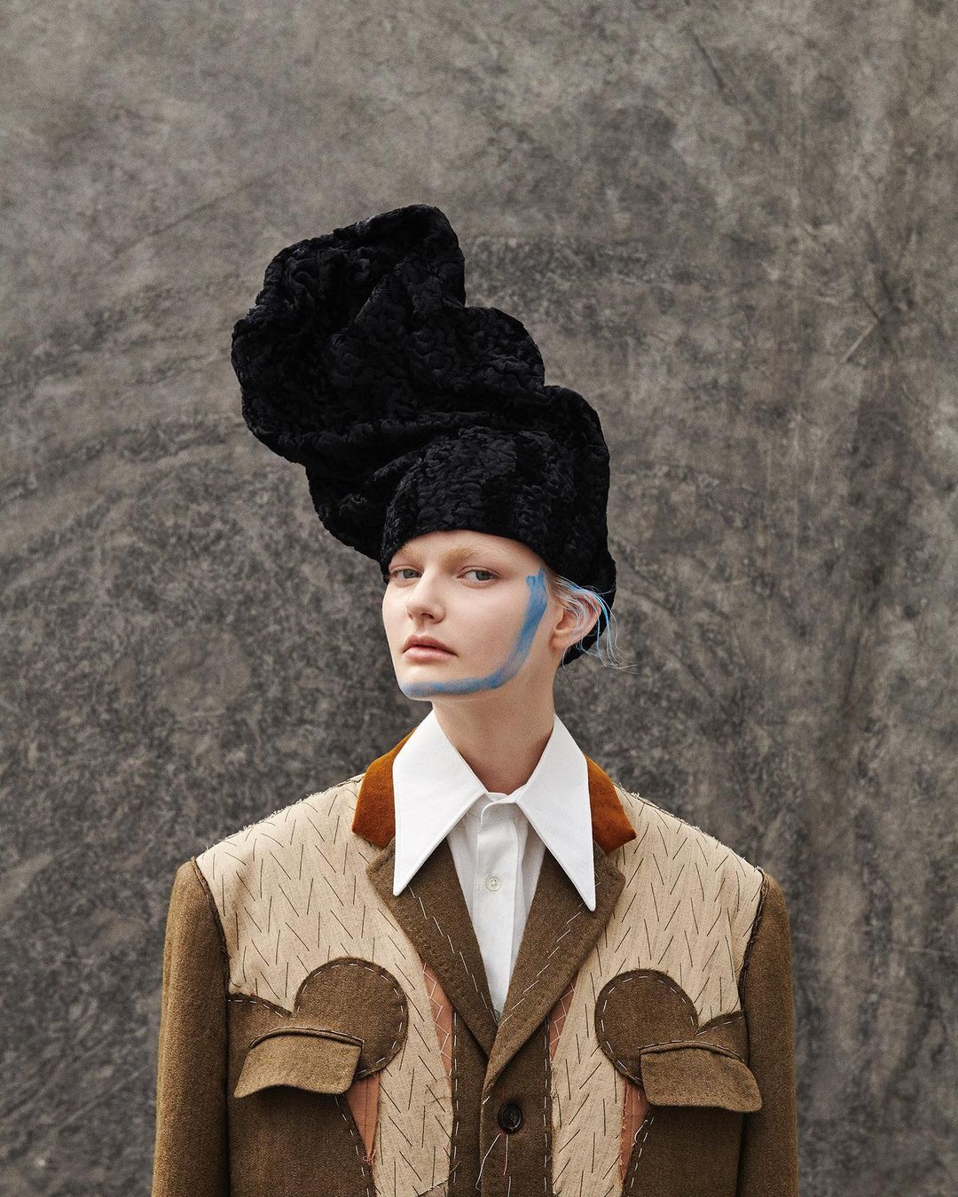 Portrait of a fair-skinned woman with blue makeup, wearing an elaborate black hat and patterned jacket against a grey mottled backdrop.