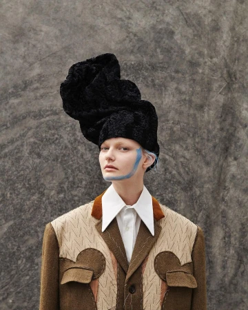 Portrait of a fair-skinned woman with blue makeup, wearing an elaborate black hat and patterned jacket against a grey mottled backdrop.