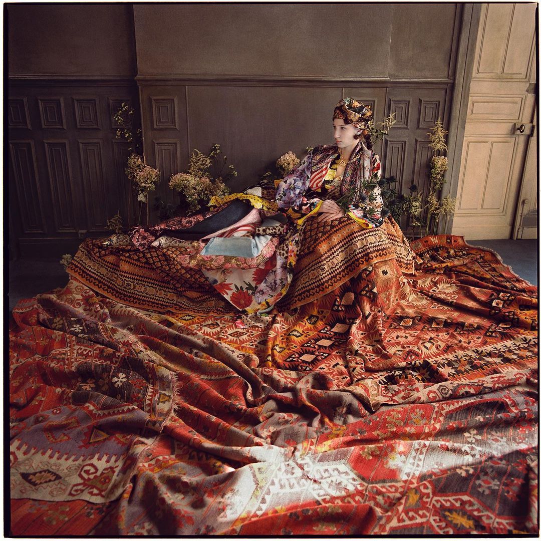 Woman in patterned headscarf and robe laying on a large patterned rug inside a room with dark brown and grey panelling on the walls.
