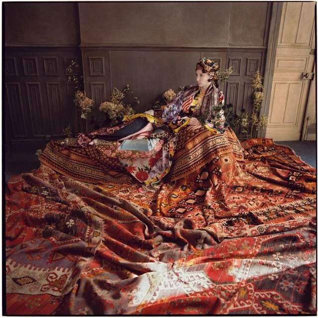 Woman in patterned headscarf and robe laying on a large patterned rug inside a room with dark brown and grey panelling on the walls.