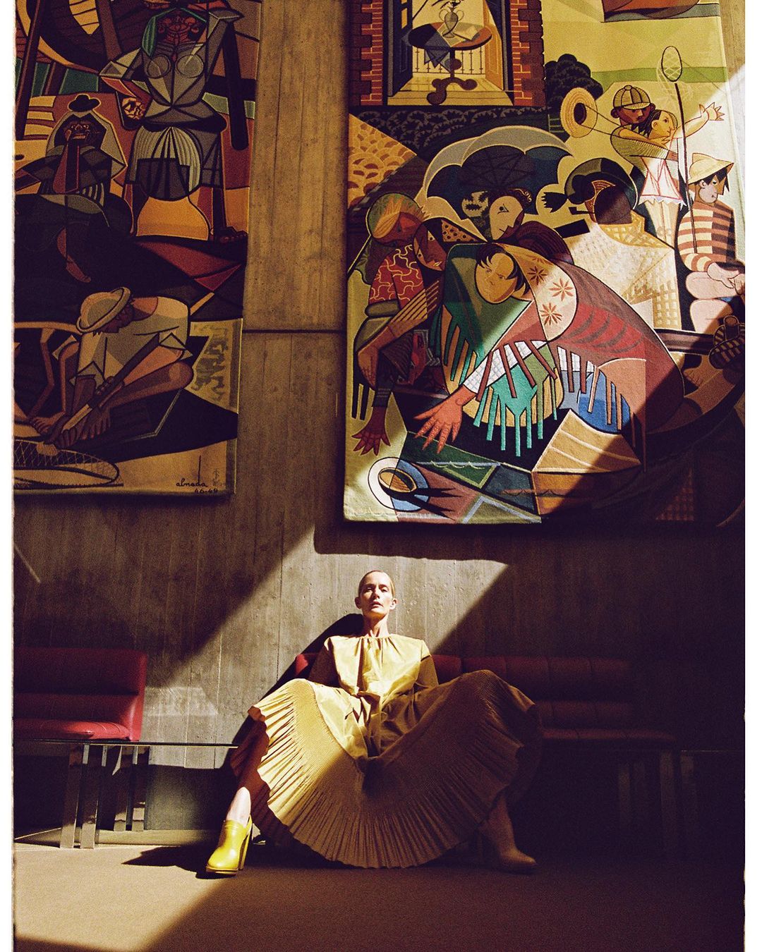 Woman in a yellow dress sits on a red bench in a room with modern art tapestries, bathed in light and shadow, creating a striking visual.