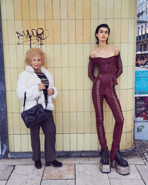 Two women stand side-by-side in an urban setting. One woman wears a fur coat and handbag, and the other wears a modern maroon outfit.