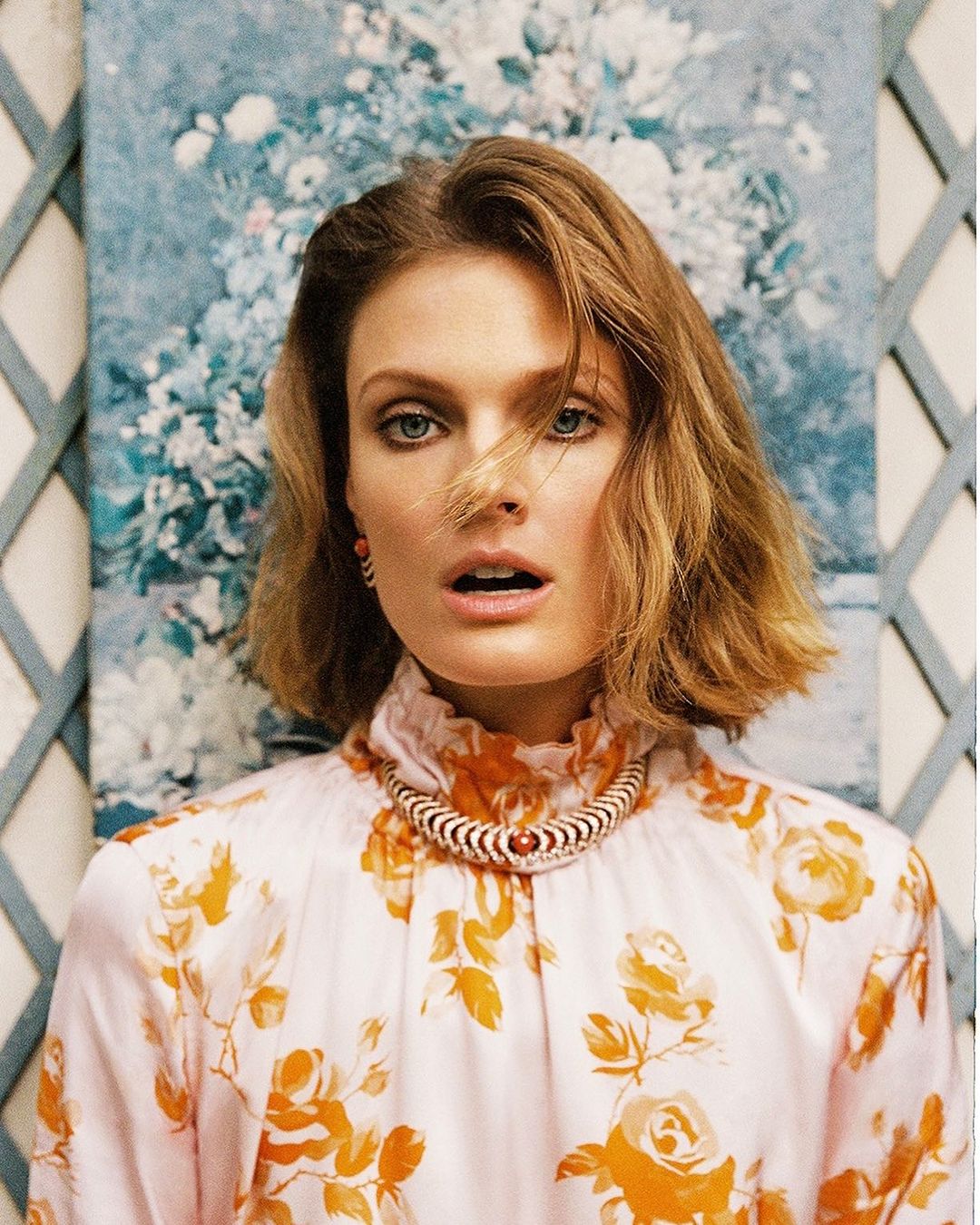 Portrait of a woman with shoulder-length blonde hair wearing a floral blouse and a necklace with a floral background in soft lighting.