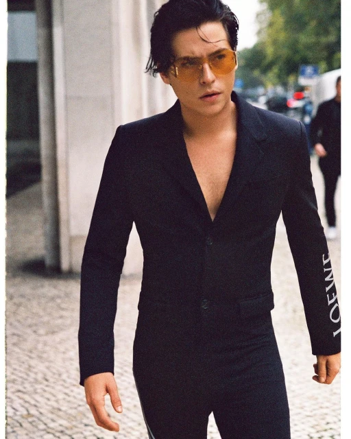 Fashion shot of a man with dark hair and yellow sunglasses wearing a black suit with white 'LOEWE' text on the sleeve outdoors.